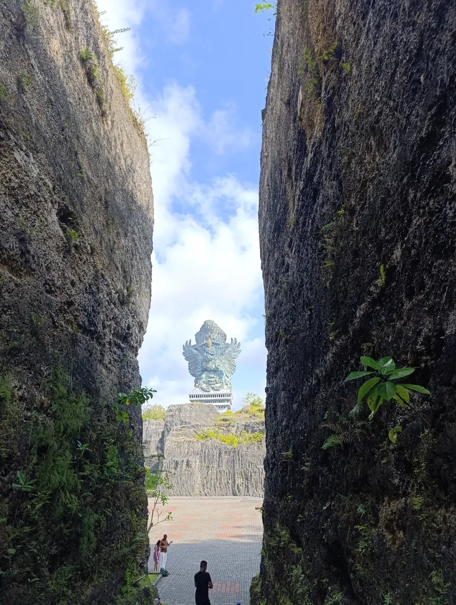 Two small figures at the end of a narrow passage between tall rock walls, looking out at a large statue in the distance